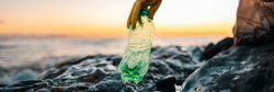 A gloved hand picks up a green plastic bottle from a rocky shoreline at sunset, with gentle ocean waves and an orange sky in the background.