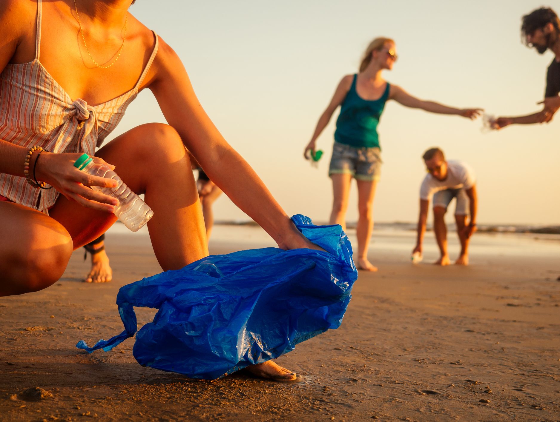 A group of people participate in a beach cleanup at sunset, with a focus on one person placing a plastic bottle into a blue bag on the sandy shore.
