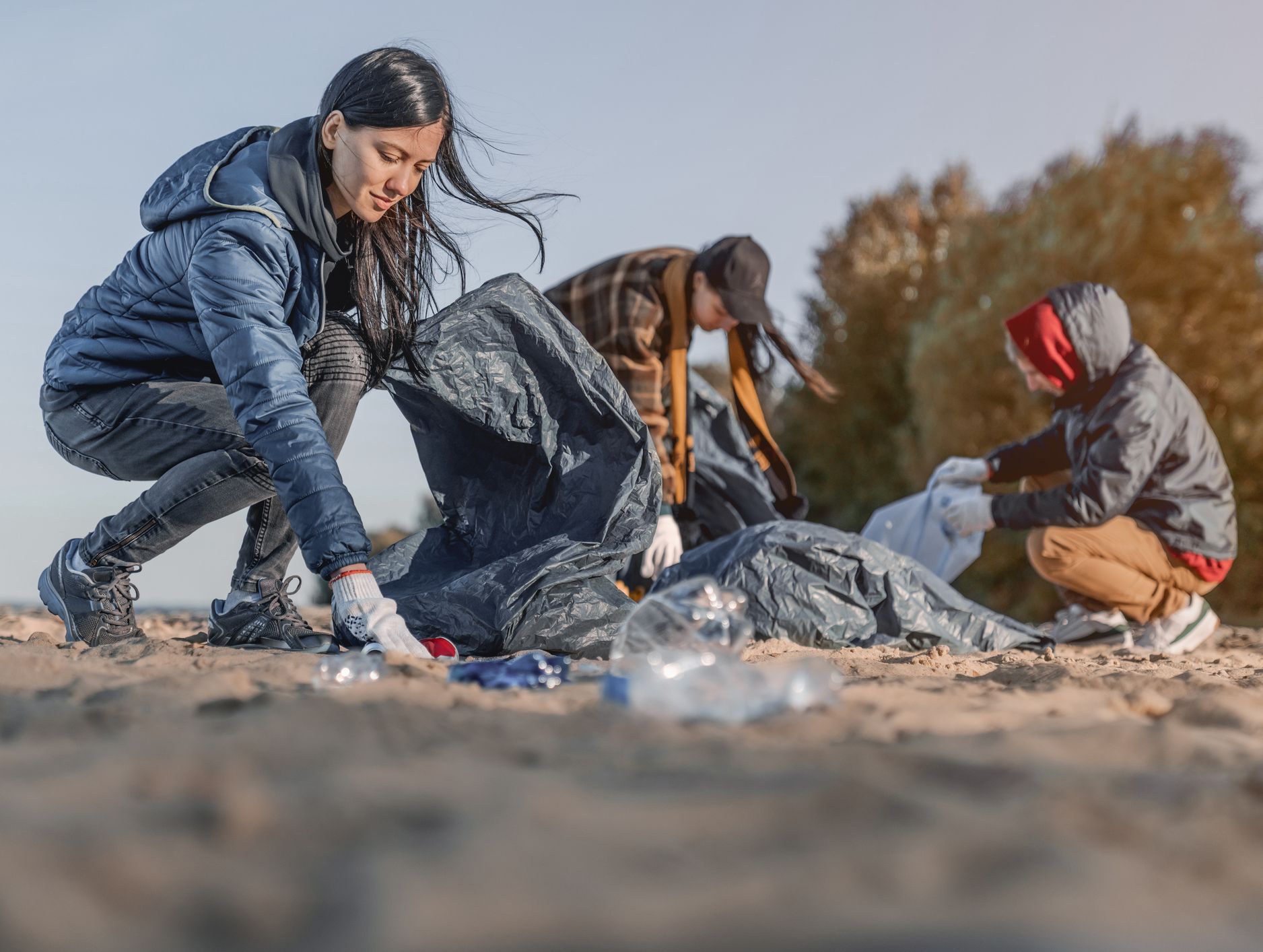 Three people in jackets and gloves collect litter on a sandy beach, placing trash into large bags, with trees and a clear sky in the background.