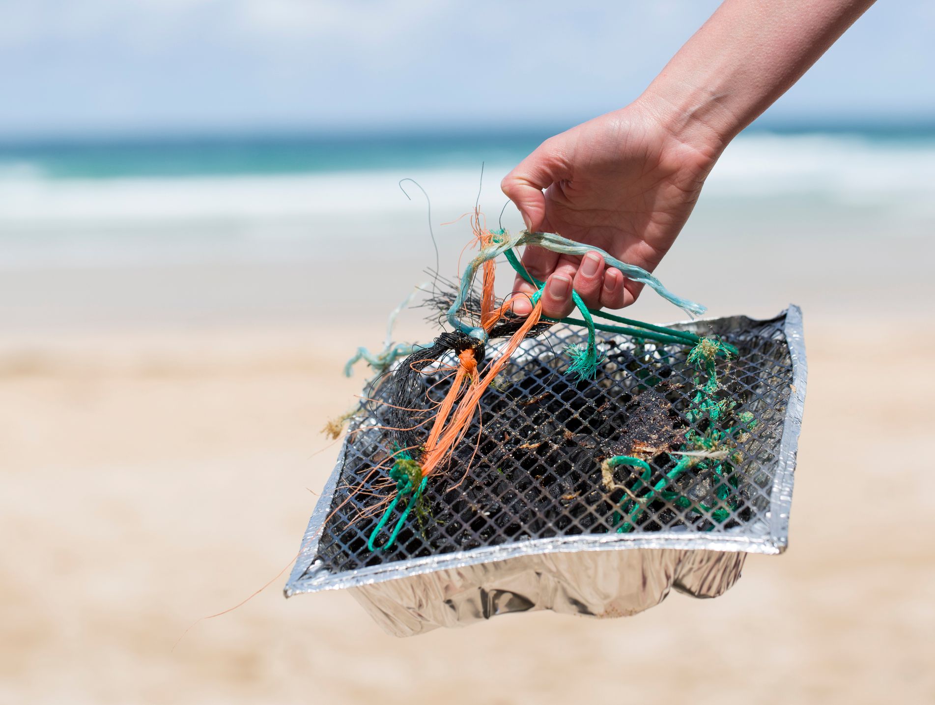 A hand holds a foil tray filled with tangled plastic debris and ropes collected from a beach, with blurred ocean waves in the background.