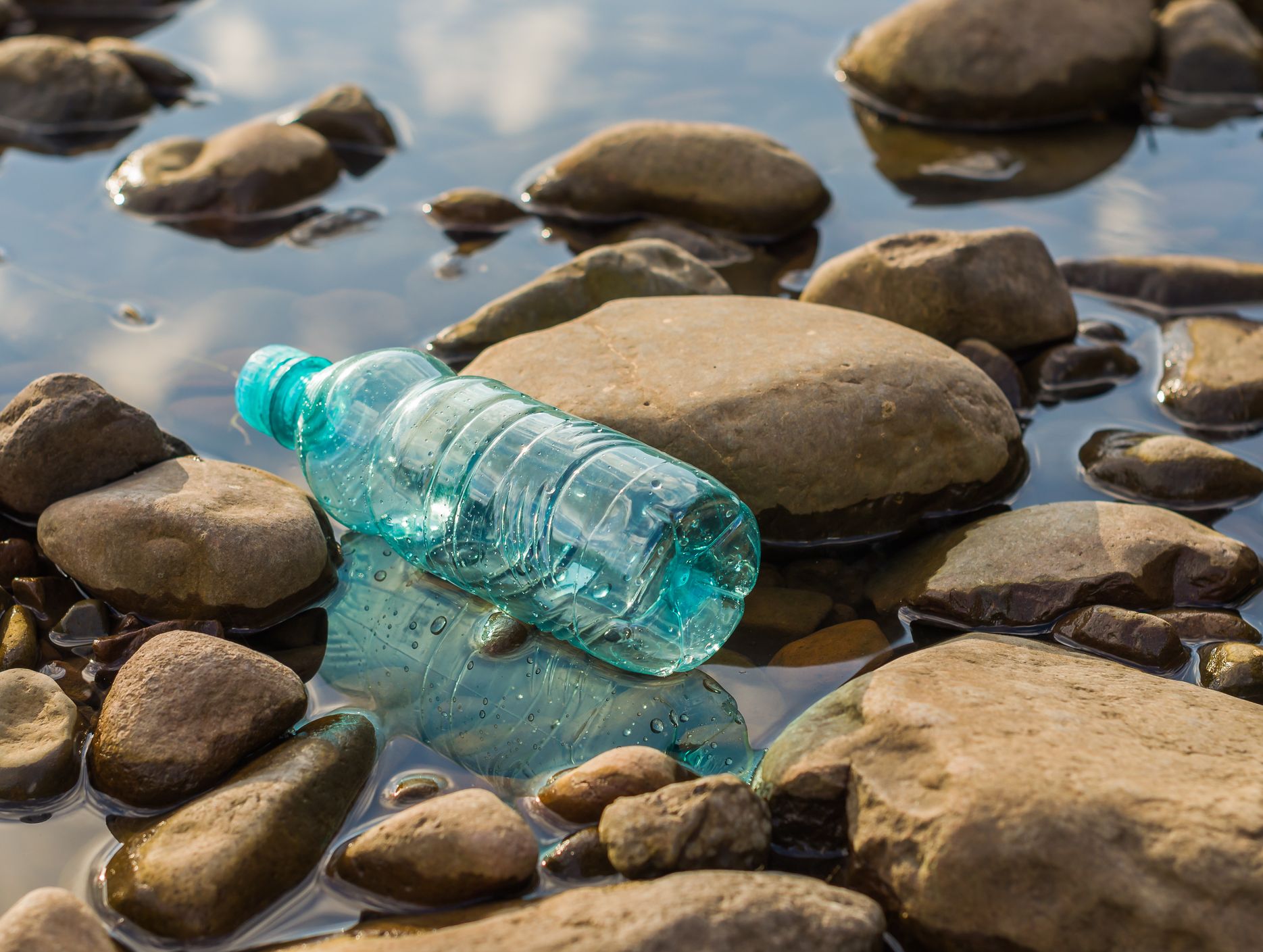 A discarded blue plastic bottle lies among smooth rocks in a shallow, reflective pool of water, highlighting pollution in a natural environment.