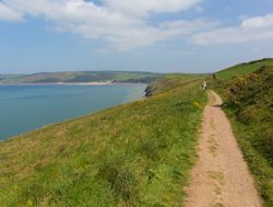 A scenic coastal trail winds along a grassy cliffside, overlooking a calm blue sea with distant rolling hills under a partly cloudy sky.
