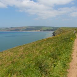 A scenic coastal trail winds along a grassy cliffside, overlooking a calm blue sea with distant rolling hills under a partly cloudy sky.