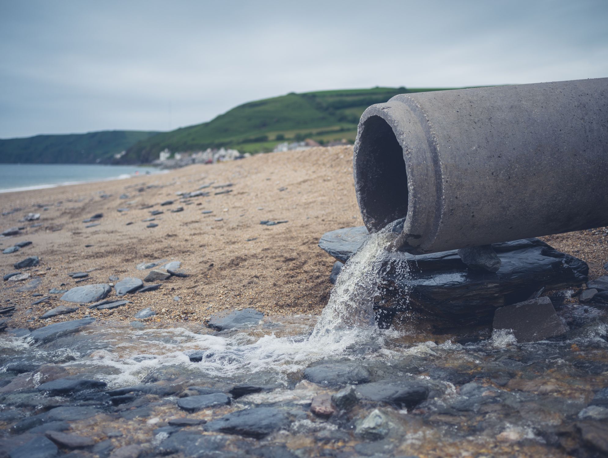 A large pipe discharges water onto a rocky beach, with hills and a distant coastal village under a cloudy sky in the background.