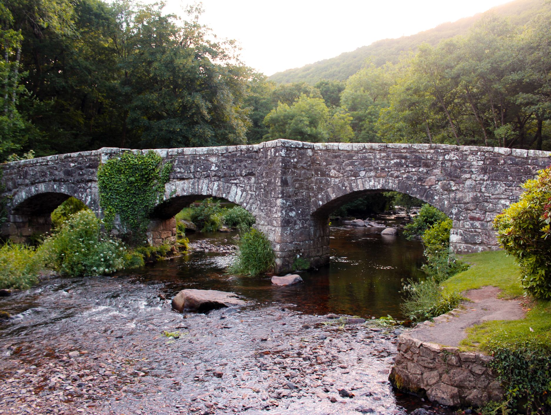 A stone bridge with ivy-covered arches spans a shallow, rocky stream surrounded by lush greenery and trees in a peaceful woodland setting.