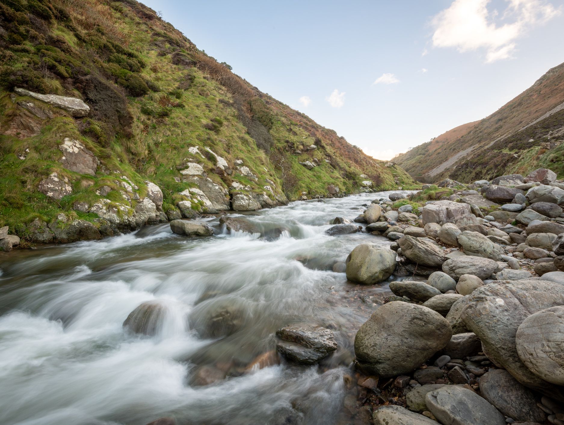 A fast-flowing river winds through a rocky valley with green, grass-covered slopes, under a clear sky with a few clouds.