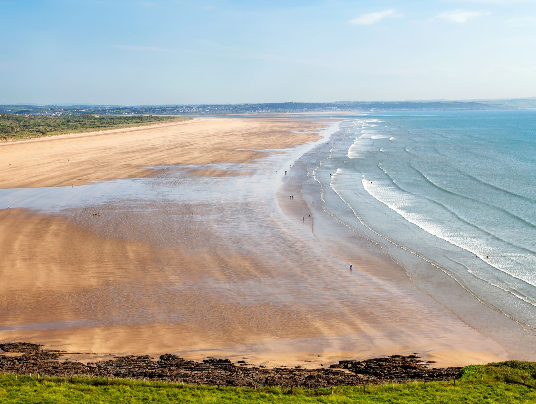 A vast sandy beach with gentle waves and scattered visitors stretches along a coastline, with distant green hills under a clear blue sky.