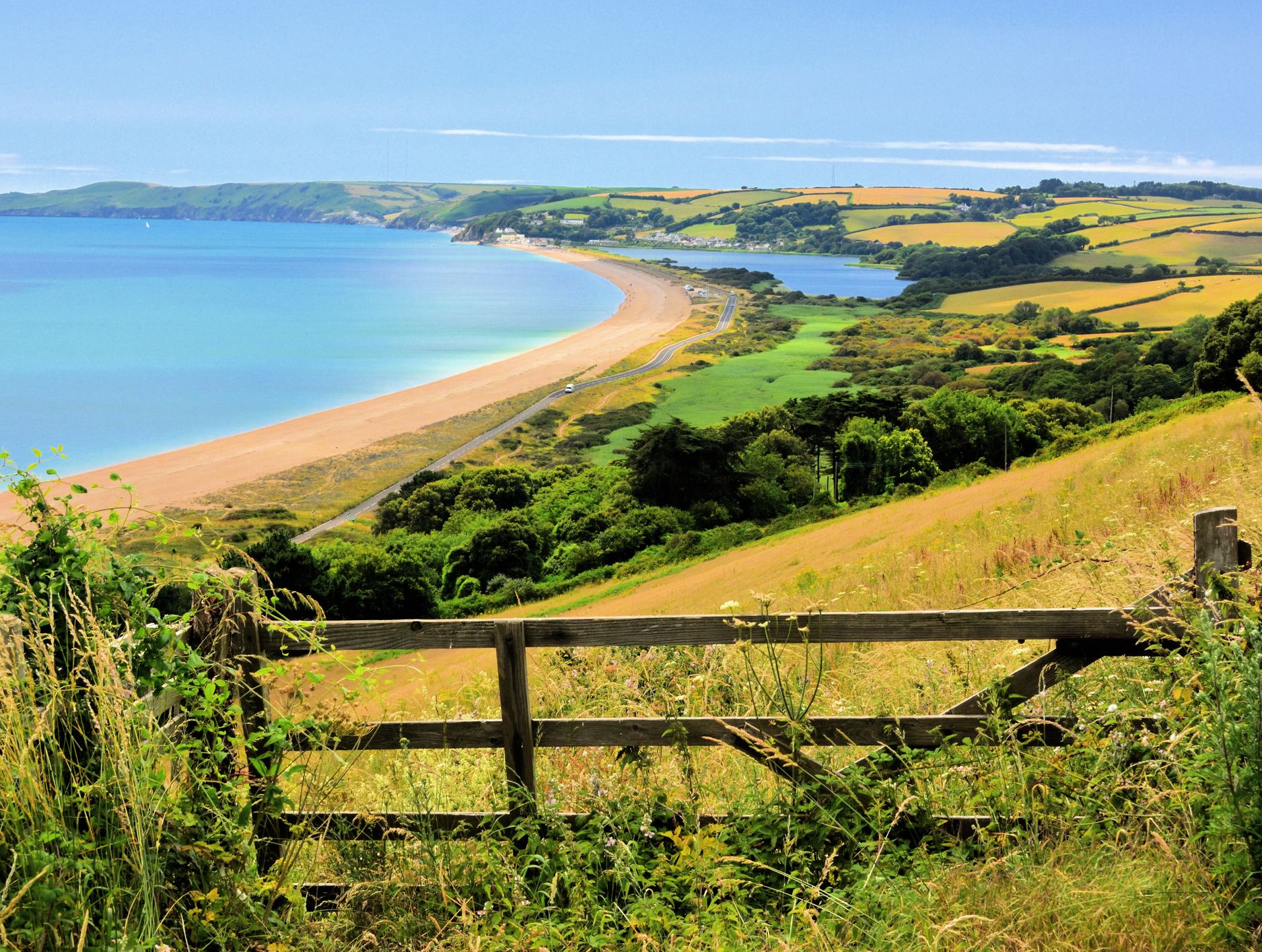 A scenic view of a curved sandy beach bordered by lush green fields and rolling hills, framed by a rustic wooden fence and wild vegetation in the foreground.