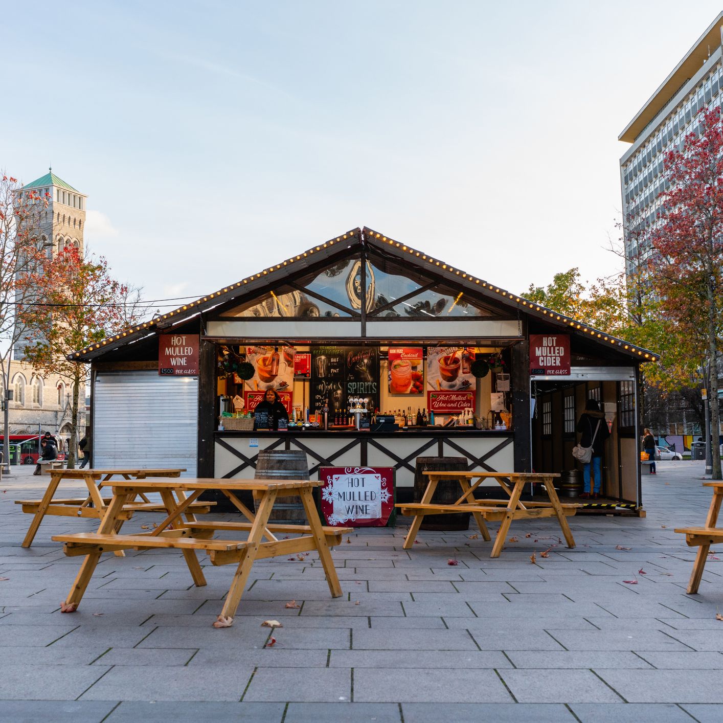 A cozy wooden stall offering hot mulled wine and cider, surrounded by picnic tables and autumn trees, at one of the charming Christmas markets in Devon.