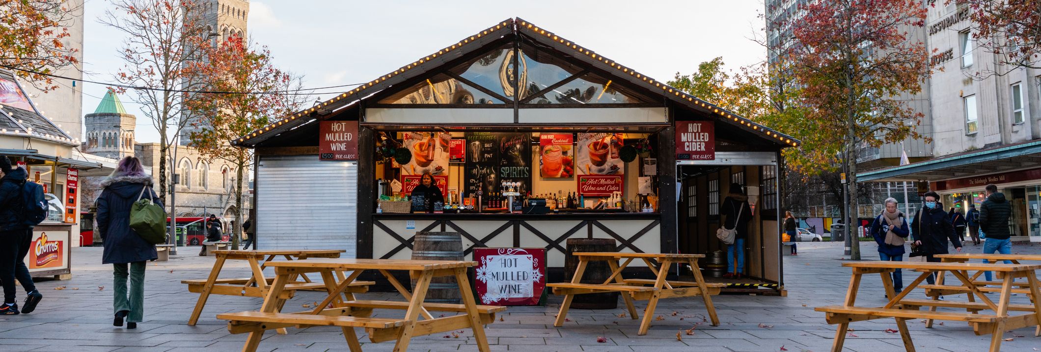 A cozy wooden stall offering hot mulled wine and cider, surrounded by picnic tables and autumn trees, at one of the charming Christmas markets in Devon.