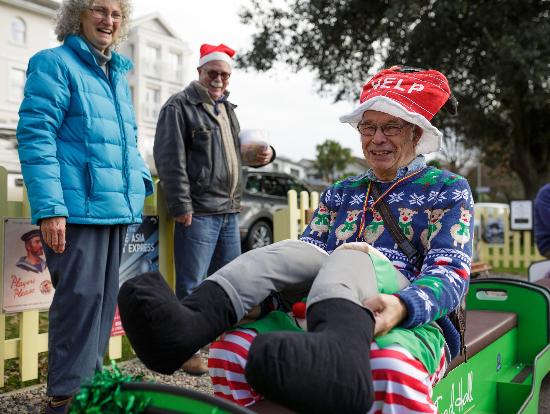 A cheerful man wearing a festive hat and Christmas sweater enjoys a playful moment on a miniature train, surrounded by smiling friends at Christmas markets in Devon.