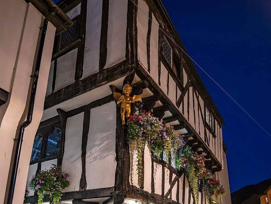 Historic Tudor-style pub in South Devon illuminated at night, adorned with vibrant hanging flower baskets and a golden cherub ornament, showcasing its charming and cozy atmosphere.