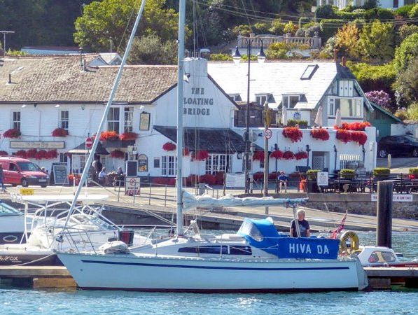 The Floating Bridge, a waterfront pub in South Devon, surrounded by scenic greenery and colorful flower displays, with yachts docked nearby adding to its picturesque coastal charm.