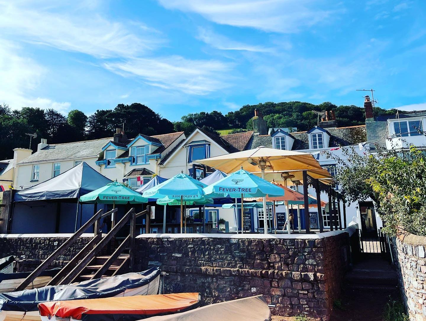 Coastal pub in South Devon featuring outdoor seating with colorful umbrellas, nestled in a hillside village under a bright blue sky, offering a relaxed and scenic atmosphere.