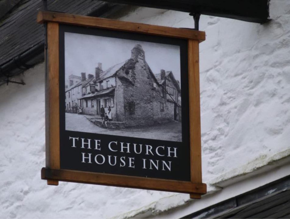 Sign for The Church House Inn, one of the historic pubs in South Devon, featuring a vintage black-and-white image of a traditional stone building, mounted on a whitewashed wall.