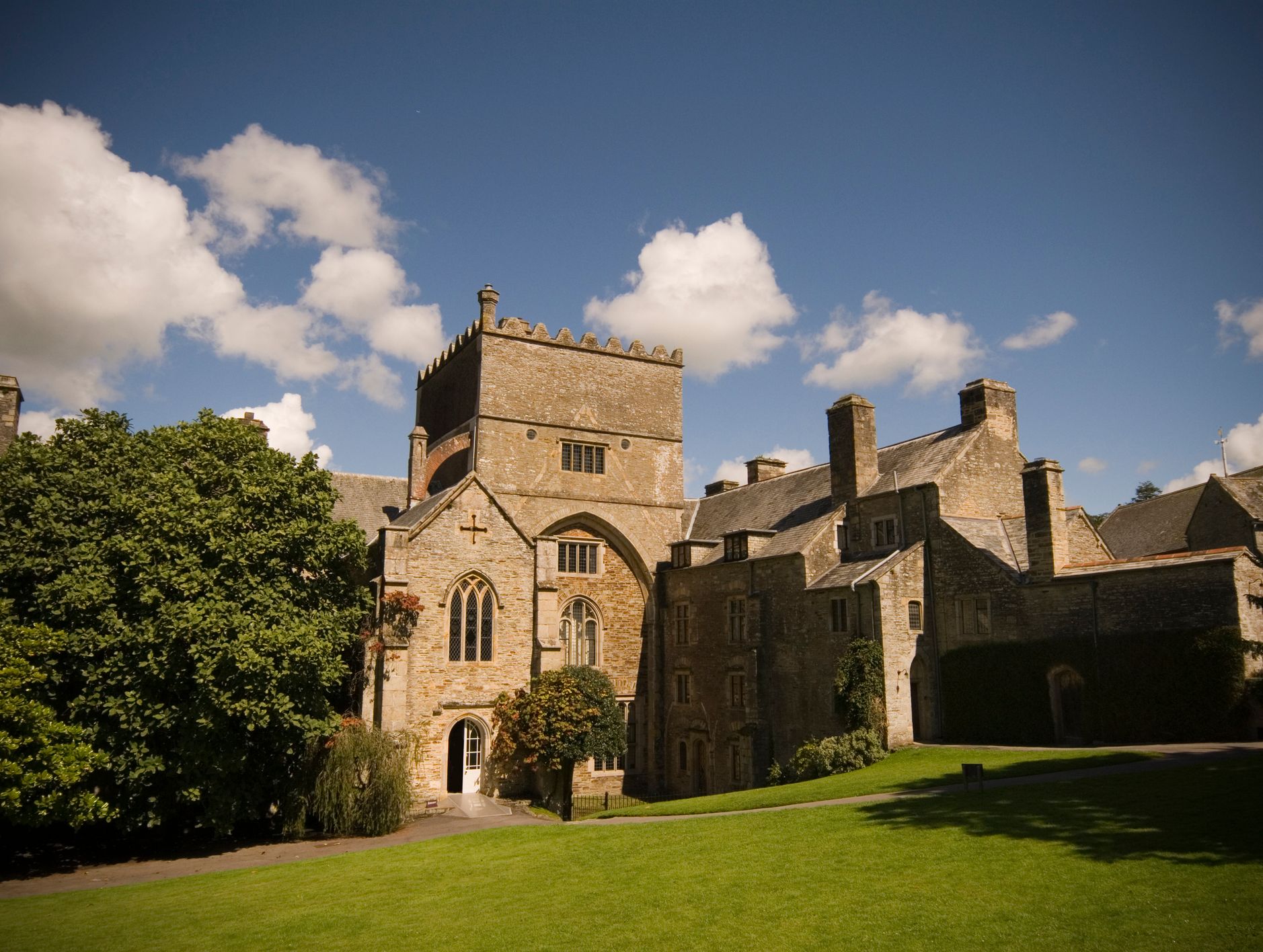 A historic stone building with Gothic architectural elements stands under a bright blue sky with scattered clouds, surrounded by green grass and lush trees.