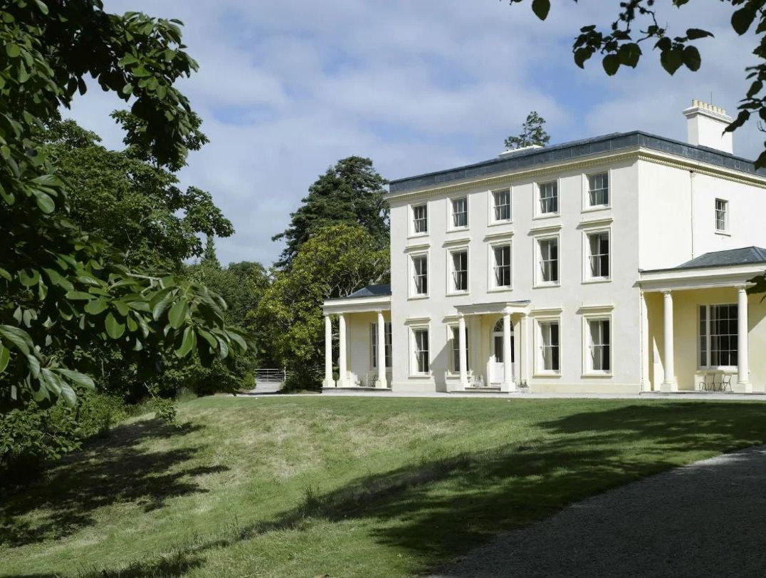 A large, elegant cream-colored Georgian-style house with tall windows and columned porches, surrounded by green lawns and trees under a partly cloudy sky.