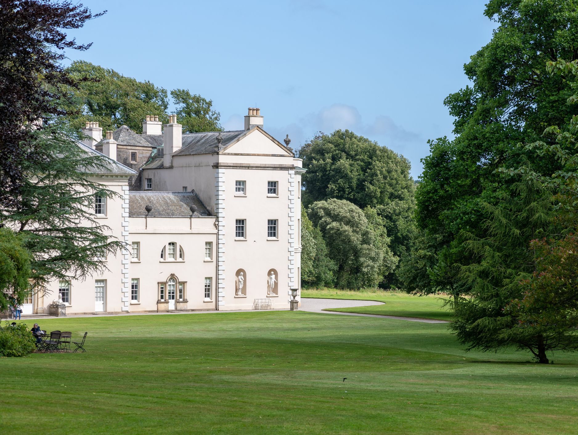 A grand cream-colored mansion with classical architecture stands surrounded by lush green lawns and tall trees under a clear blue sky.
