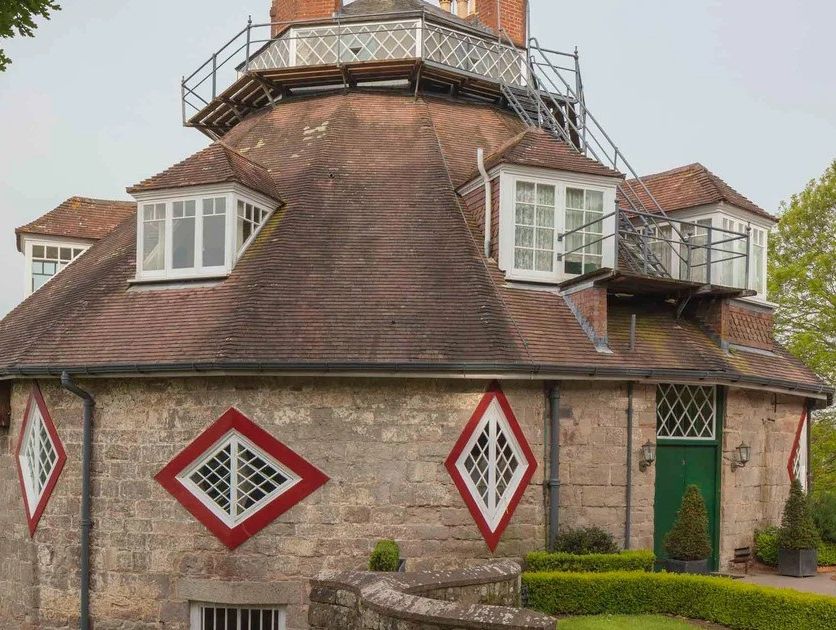 A unique round stone house with red diamond-shaped windows, a brown tiled roof, and green doors, surrounded by a garden with potted plants and lush greenery.