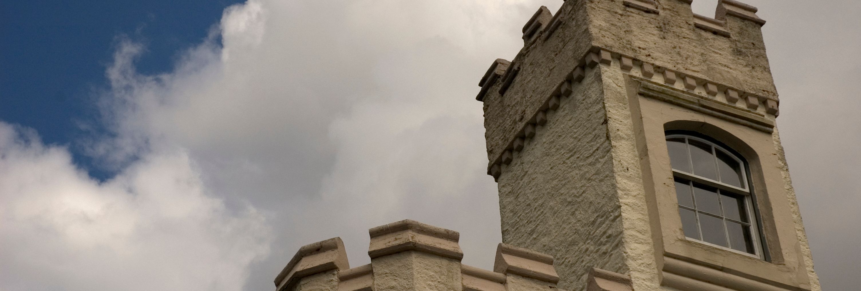 A close-up view of a cream-colored castle tower with battlements and an arched window, set against a partly cloudy sky with large, billowing clouds.
