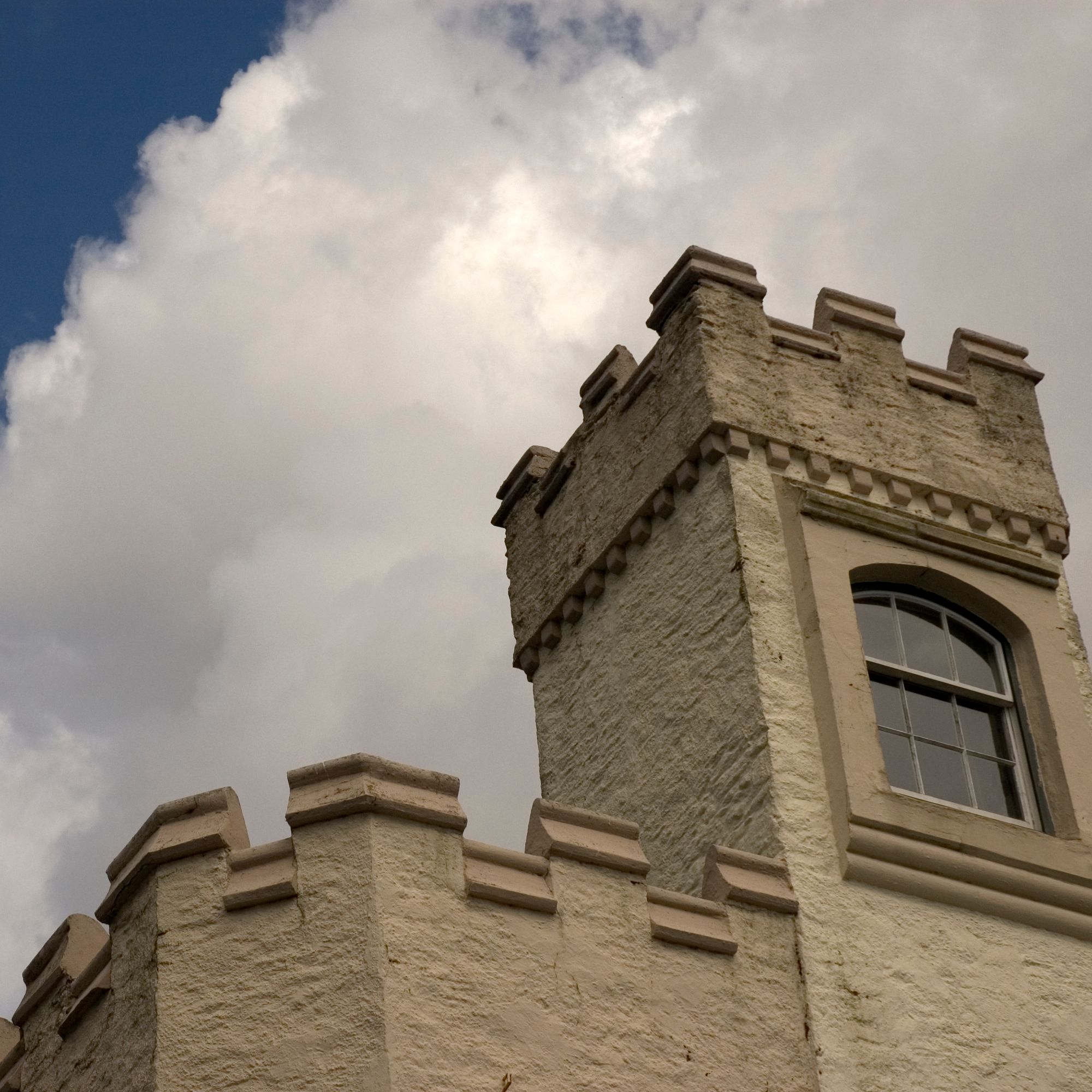 A close-up view of a cream-colored castle tower with battlements and an arched window, set against a partly cloudy sky with large, billowing clouds.