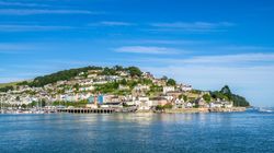 A scenic view of a coastal town with colorful houses on a hillside, surrounded by greenery, overlooking a calm harbor filled with sailboats, under a clear blue sky.