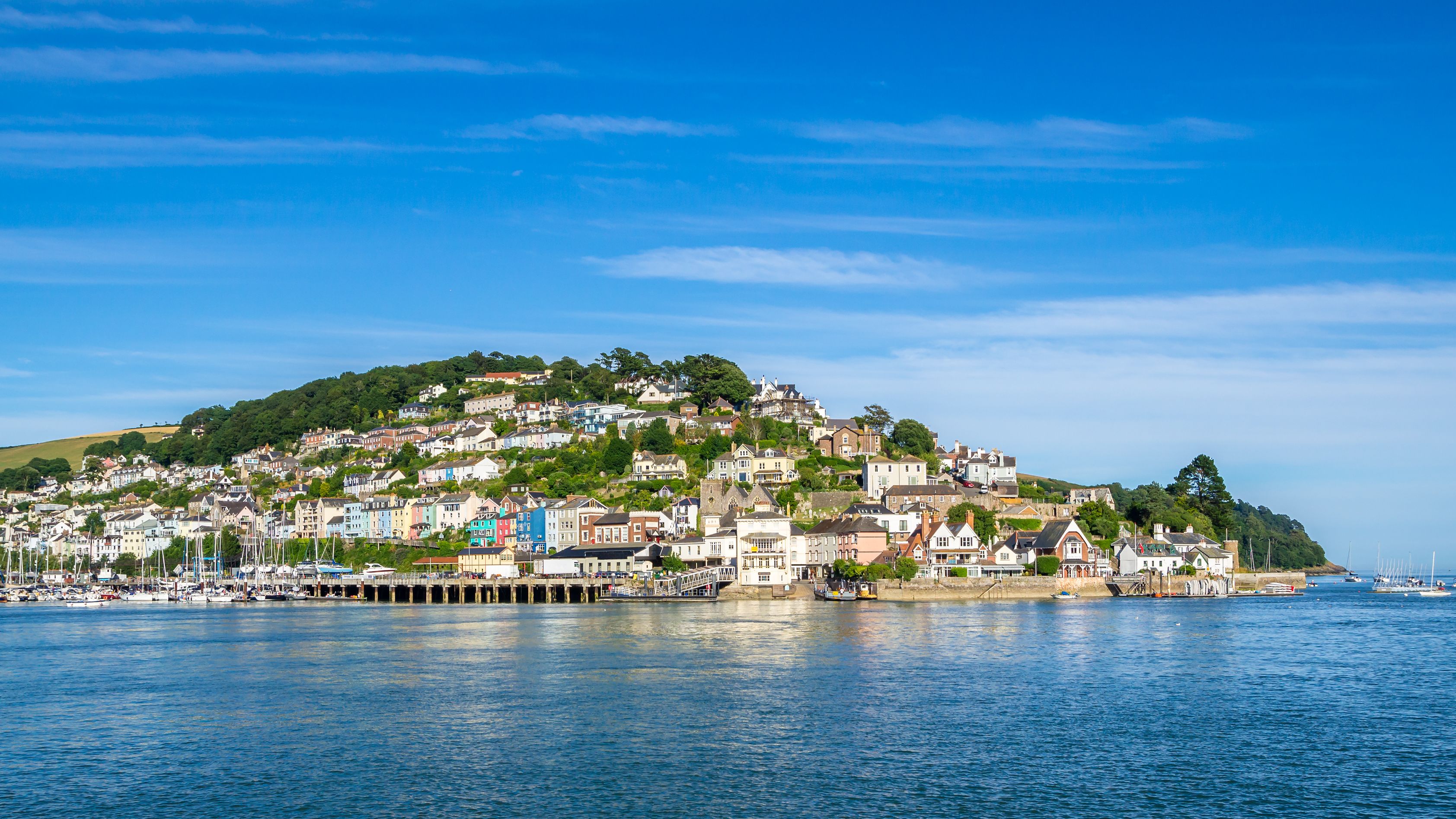 A scenic view of a coastal town with colorful houses on a hillside, surrounded by greenery, overlooking a calm harbor filled with sailboats, under a clear blue sky.