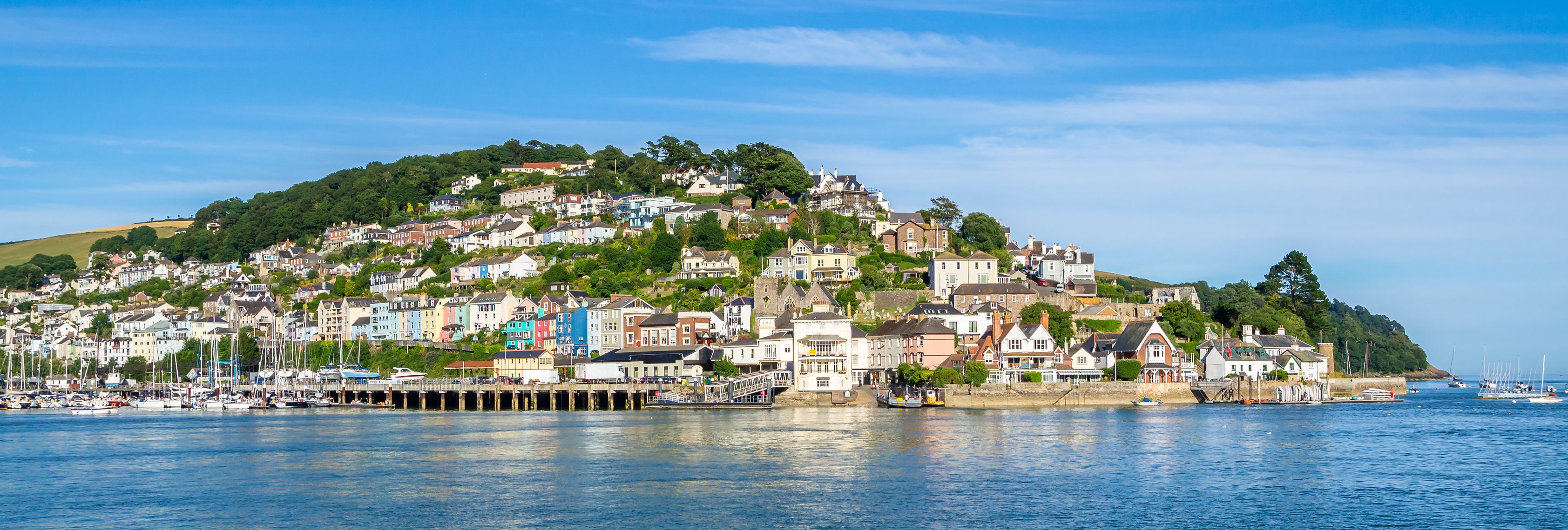A scenic view of a coastal town with colorful houses on a hillside, surrounded by greenery, overlooking a calm harbor filled with sailboats, under a clear blue sky.