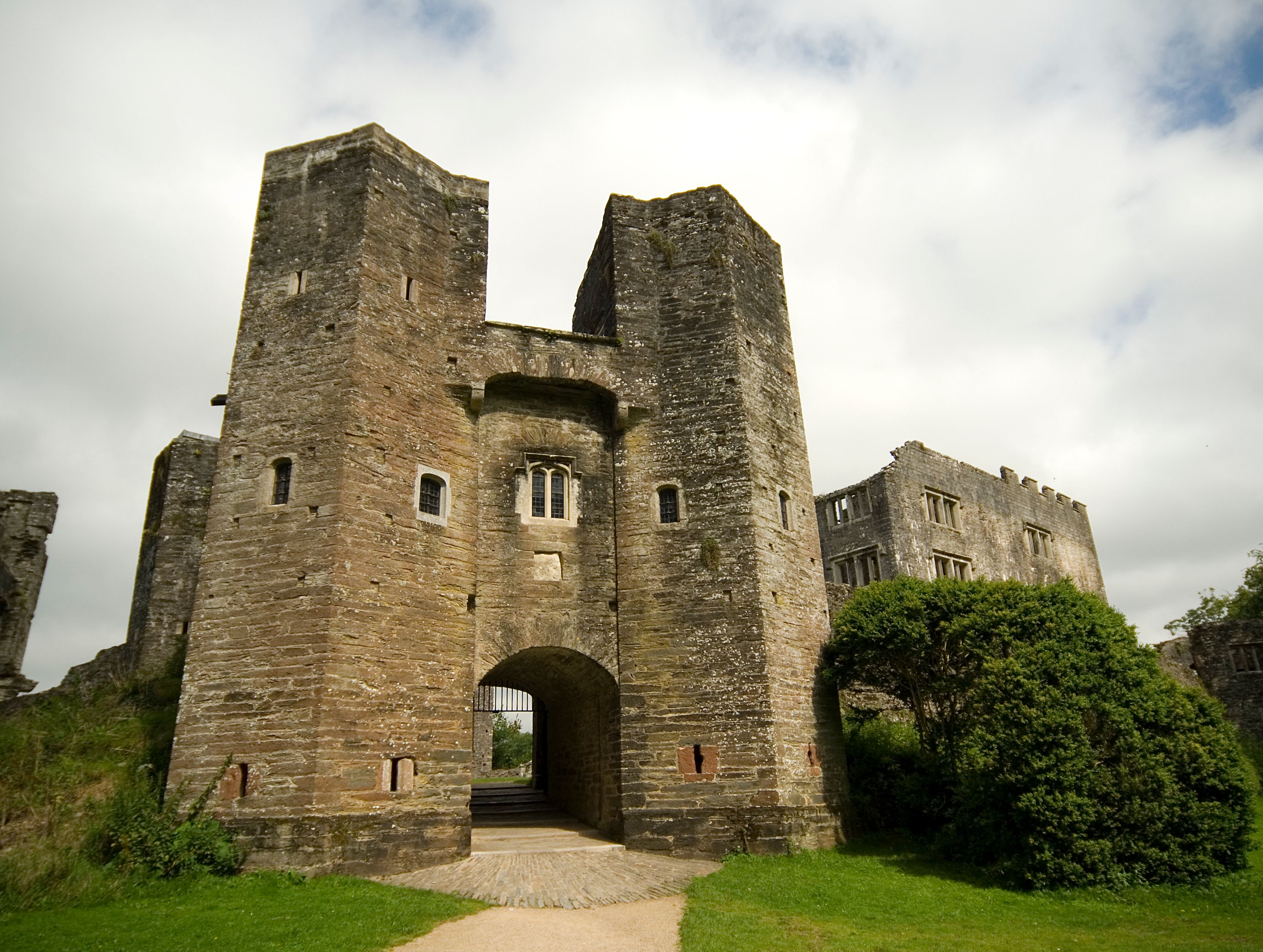 A historic stone castle entrance with tall towers and arched gate, surrounded by green grass and trees, under a partly cloudy sky.