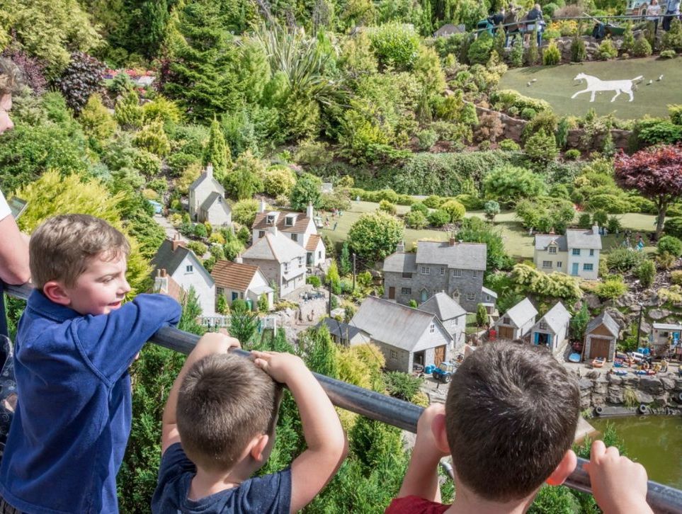 Children and an adult look over a railing at a detailed miniature village surrounded by lush greenery, with a hillside displaying a white horse figure in the background.