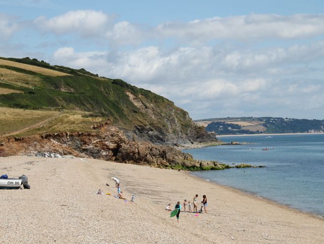 A scenic pebble beach with scattered visitors, small boats onshore, and grassy cliffs extending into the distance, beside calm blue waters under a partly cloudy sky.