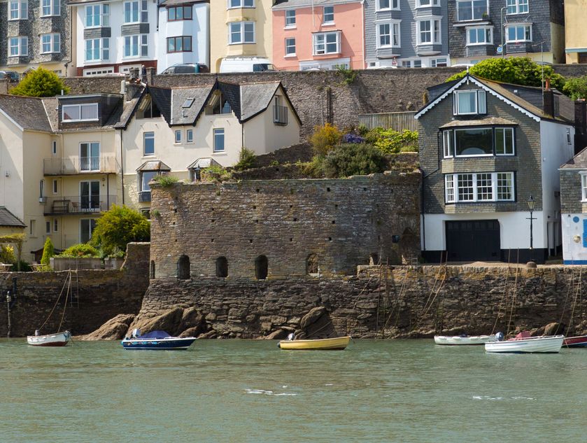 A vibrant hillside town with colorful houses overlooking a calm river, surrounded by lush greenery, with small boats moored along the stone waterfront.