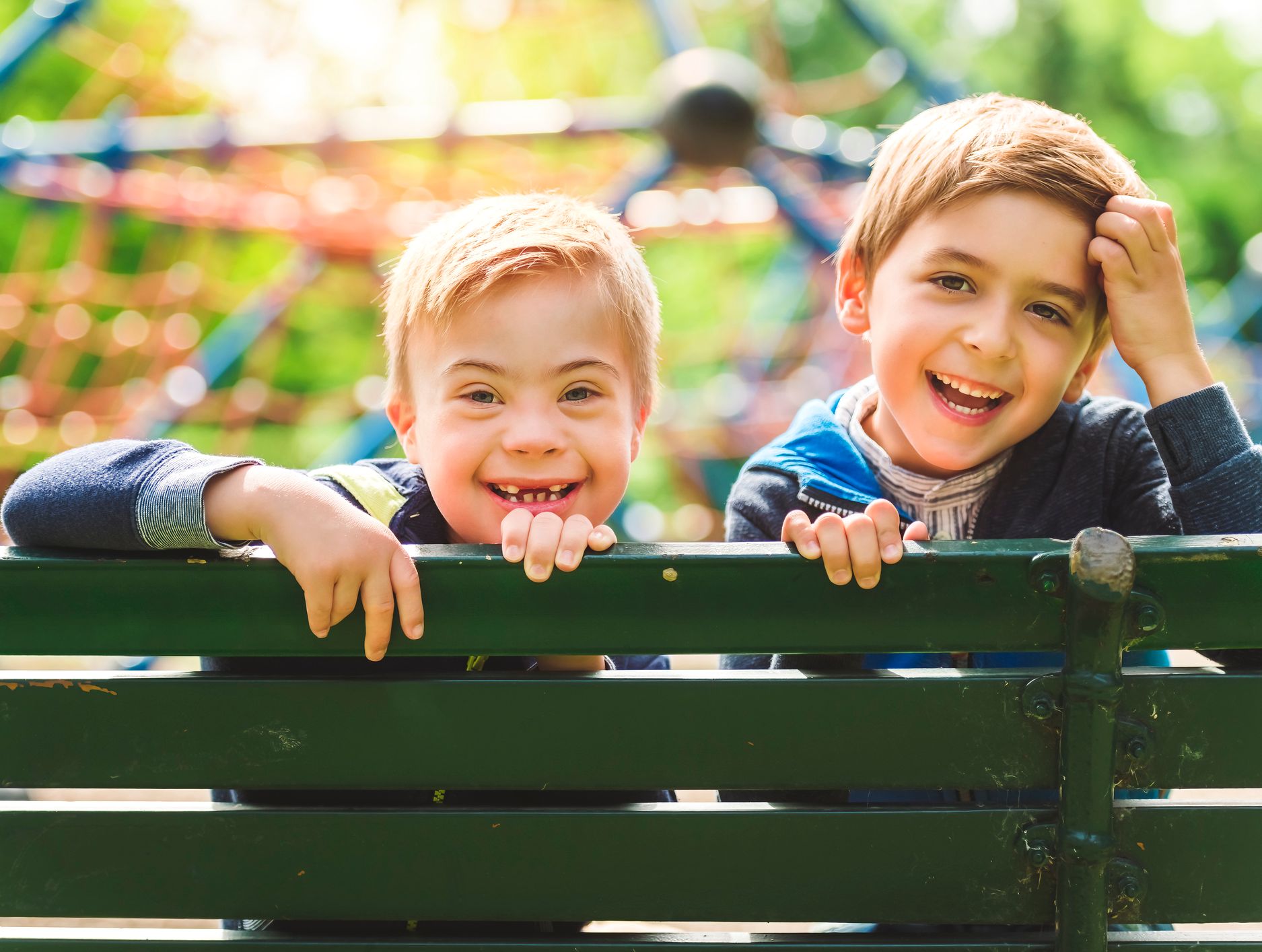 Two young boys smiling brightly over the back of a green bench at a sunny playground, with colorful climbing equipment and blurred greenery in the background.