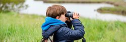 A young boy wearing a blue jacket and camouflage backpack uses binoculars to watch a flock of flamingos flying over a marshy landscape with tall grass and water.