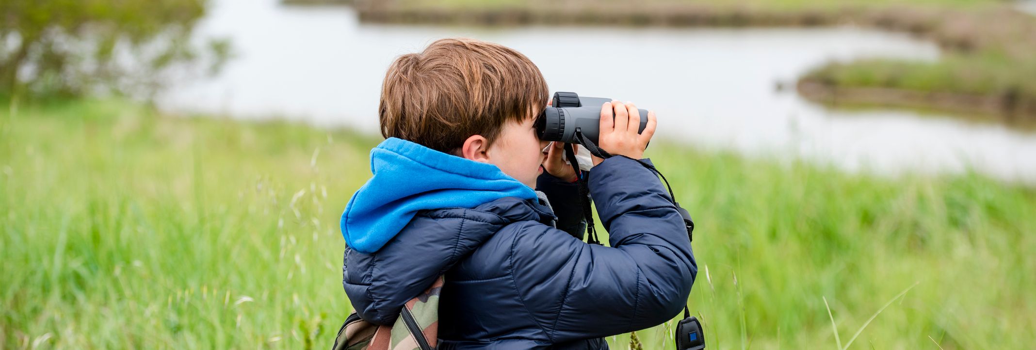 A young boy wearing a blue jacket and camouflage backpack uses binoculars to watch a flock of flamingos flying over a marshy landscape with tall grass and water.