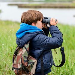 A young boy wearing a blue jacket and camouflage backpack uses binoculars to watch a flock of flamingos flying over a marshy landscape with tall grass and water.