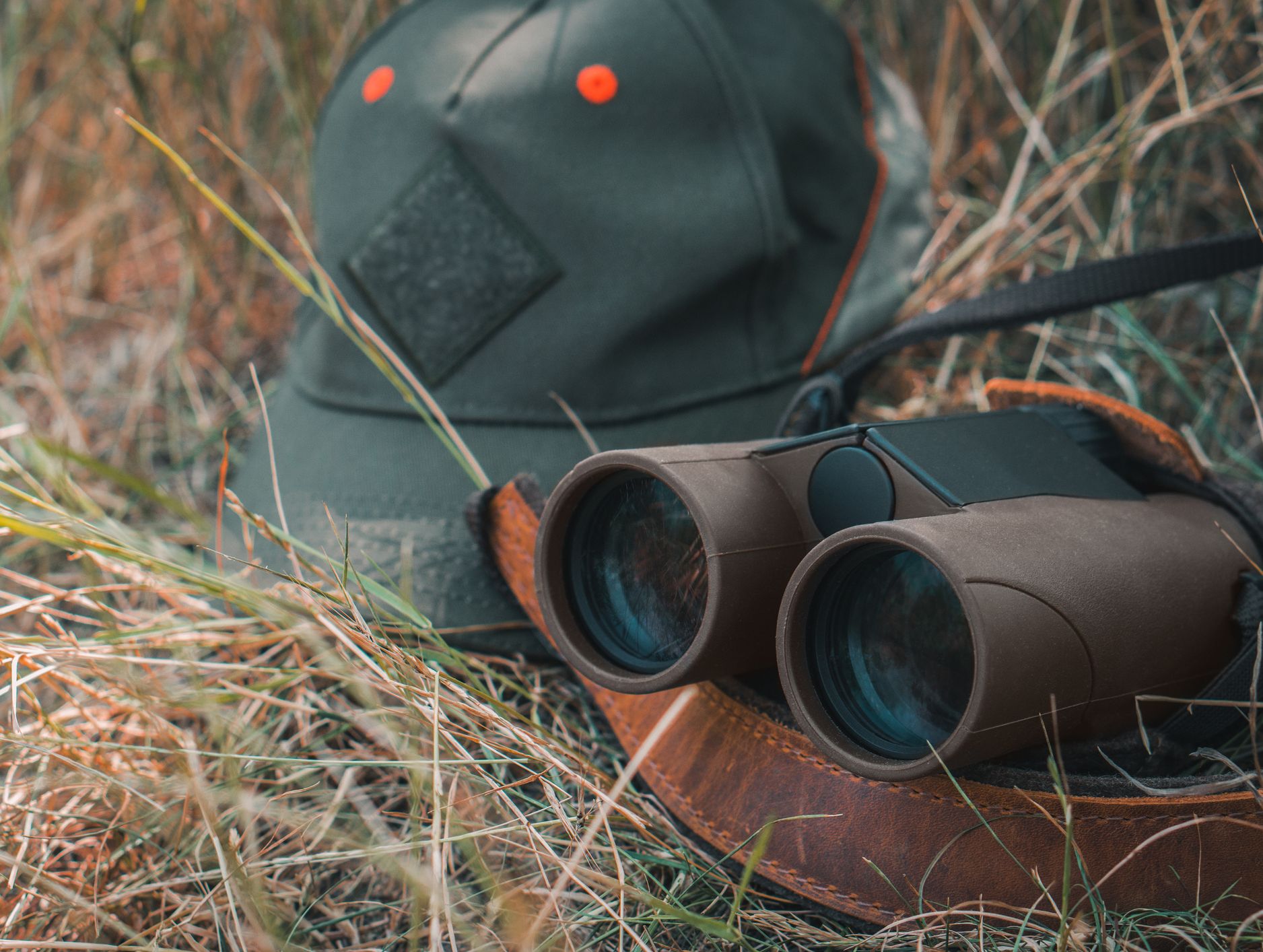 A pair of binoculars resting on a leather strap next to a green cap with orange accents, placed in dry grass under soft, natural lighting.