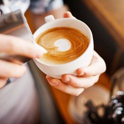 A barista pours steamed milk into a cup of coffee, creating delicate latte art. The warm café setting features a wooden counter and neatly stacked saucers in the background.