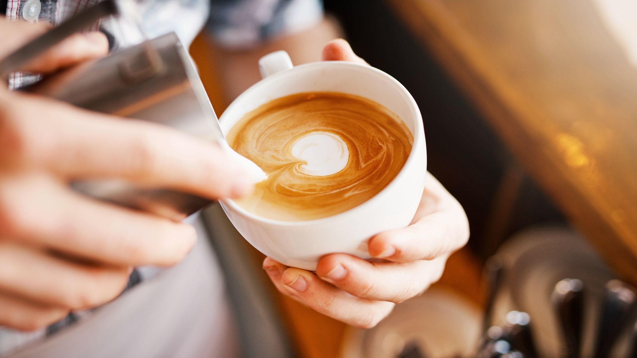 A barista pours steamed milk into a cup of coffee, creating delicate latte art. The warm café setting features a wooden counter and neatly stacked saucers in the background.