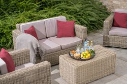Outdoor wicker patio furniture set with beige cushions and red pillows, featuring a table with drinks, lemons, and snacks.