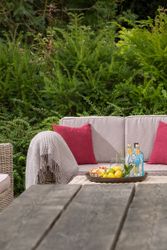 Outdoor seating area with beige sofa, red pillows, and a tray of drinks and fruit on a table, surrounded by green bushes.