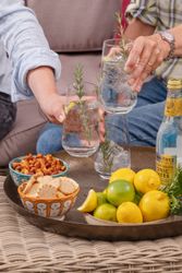Two people holding glasses with clear drinks, garnished with rosemary, with snacks and lemons on a tray