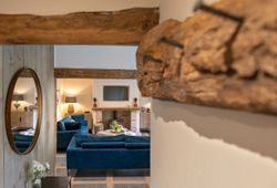 Cozy living room with exposed wooden beams, plush blue sofas, a fireplace, and a round mirror on the wall.