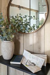 Decorative scene with a round mirror, a vase of greenery, a book, a map, and reading glasses on a shelf.