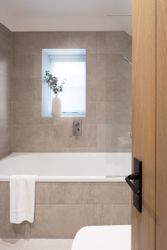 Modern bathroom with a bathtub, stone tile walls, a small window with a white vase of greenery, and a towel hanging on the tub.