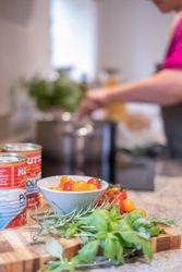 Fresh herbs, cherry tomatoes, and canned tomatoes on a kitchen counter with a person cooking in the background