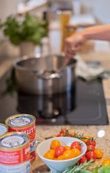 Person stirring a pot on a stovetop with canned tomatoes, cherry tomatoes, and herbs on the counter.