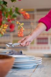 Person reaching for silverware beside a stack of white plates on a table with flowers in the foreground.