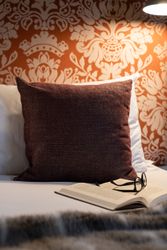 A cozy bed with a large pillow, an open book, and a pair of glasses under a reading lamp.