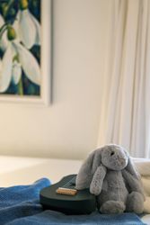 Stuffed grey bunny sitting next to a small guitar on a bed with a blue blanket.